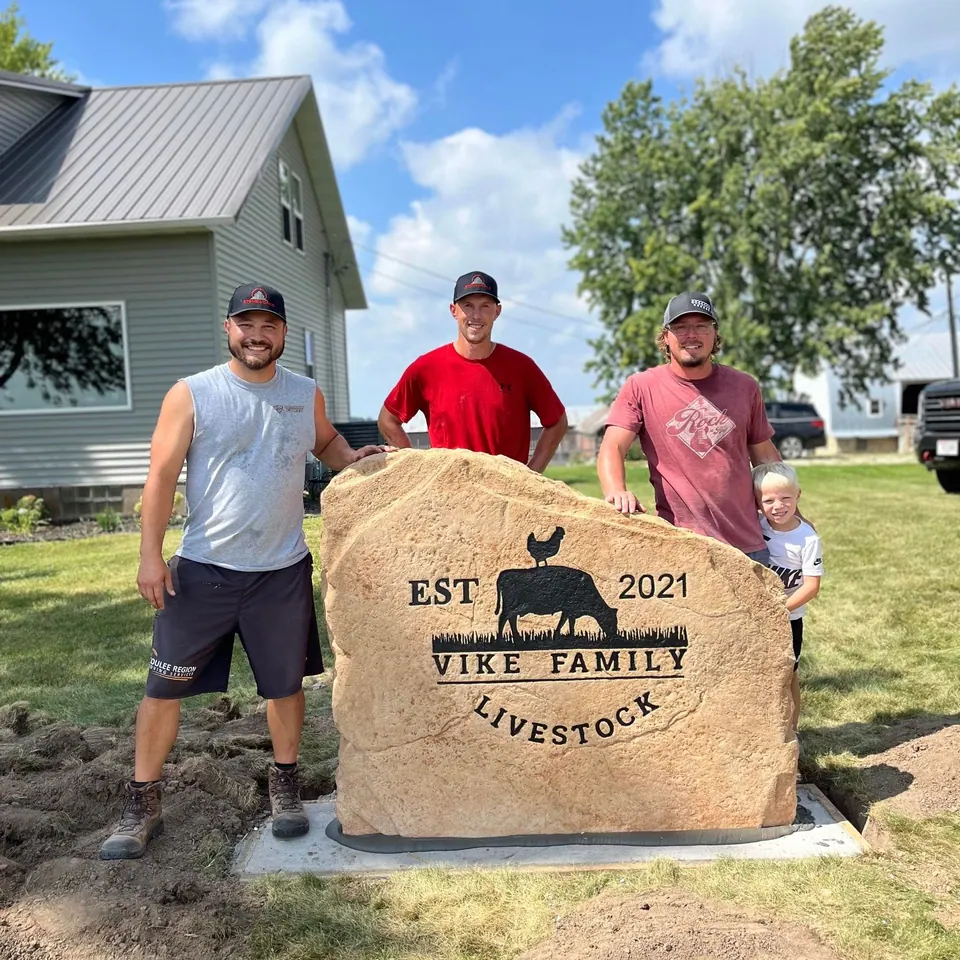 Large engraved farm entrance boulder with a livestock logo, three men and a child standing beside it for scale