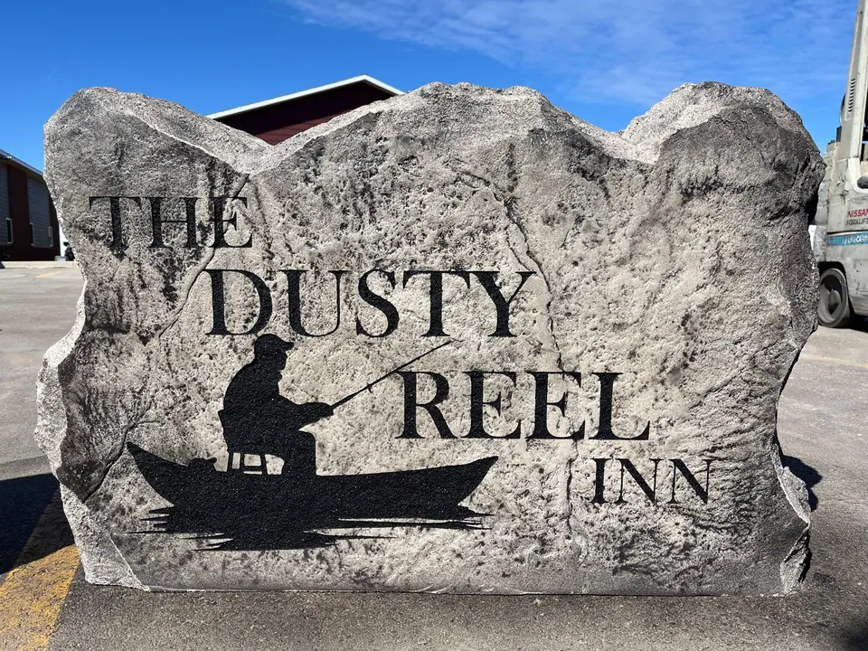 Grey inn and restaurant boulder sign with large lettering and a fisherman-in-a-boat silhouette